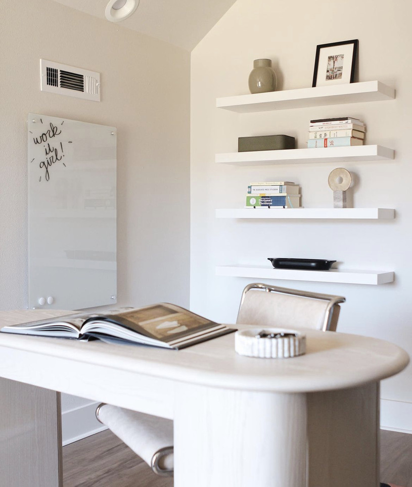 Modern desk with book open on top. Wall-mounted white board with Work It Girl Text. Floating white shelving with decor and books. 