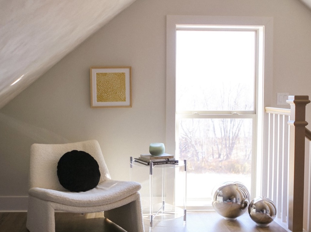 Modern sitting area with window looking out at woods and sunshine. @ silver floor orbs, white chair, black round pillow and side table with books and candle. Small yellow art print on wall.